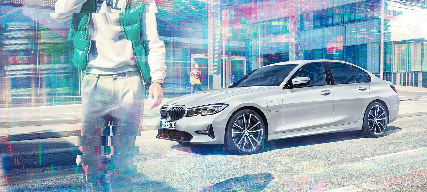 BMW 3 серии Седан, боковой снимок BMW 330е. Боковой снимок стоящего BMW 330e iPerformance Sedan перед городским стеклянным зданием с идущими пассажирами, три четверти.