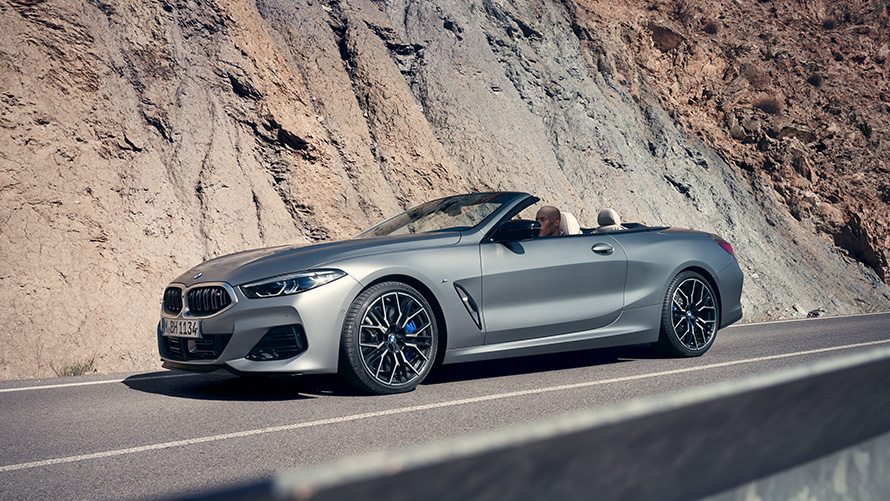 BMW 8 серии Cabrio G14 LCI (рестайлинг 2022 года) с убранным мягким верхом в движении на фоне отвесной скалы BMW 8 серии Cabrio G14 LCI (рестайлинг 2022 года) эксклюзивного цвета BMW Individual Морозный Серый металлик с убранным мягким верхом в движении на фоне отвесной скалы, вид сбоку