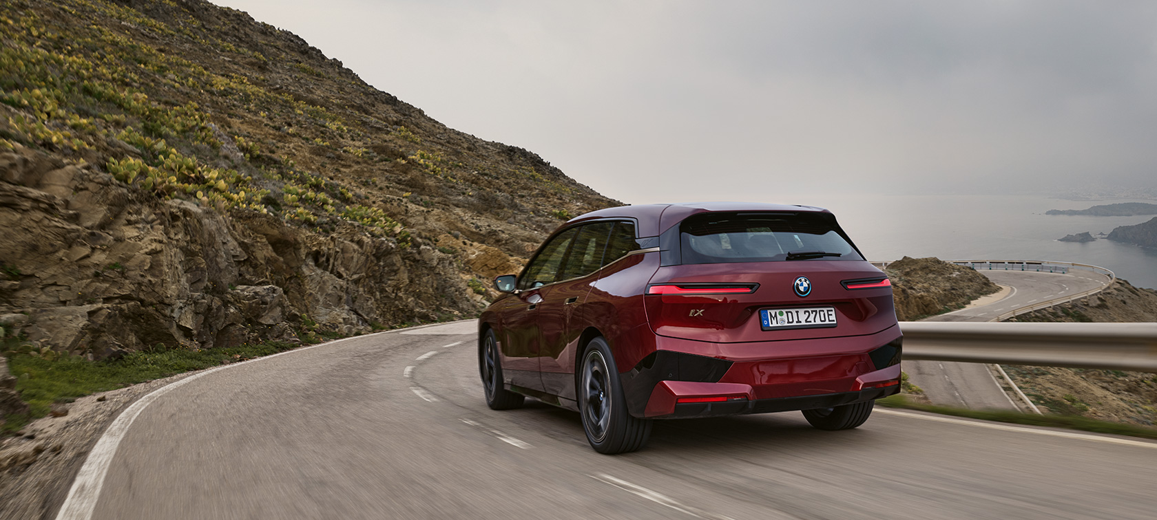 Электрический SUV BMW iX i20 2021 xDrive50, вид сзади в движении по набережной Электрический SUV BMW iX i20 2021 xDrive50 цвета Aventurin Red, вид сзади в движении по набережной