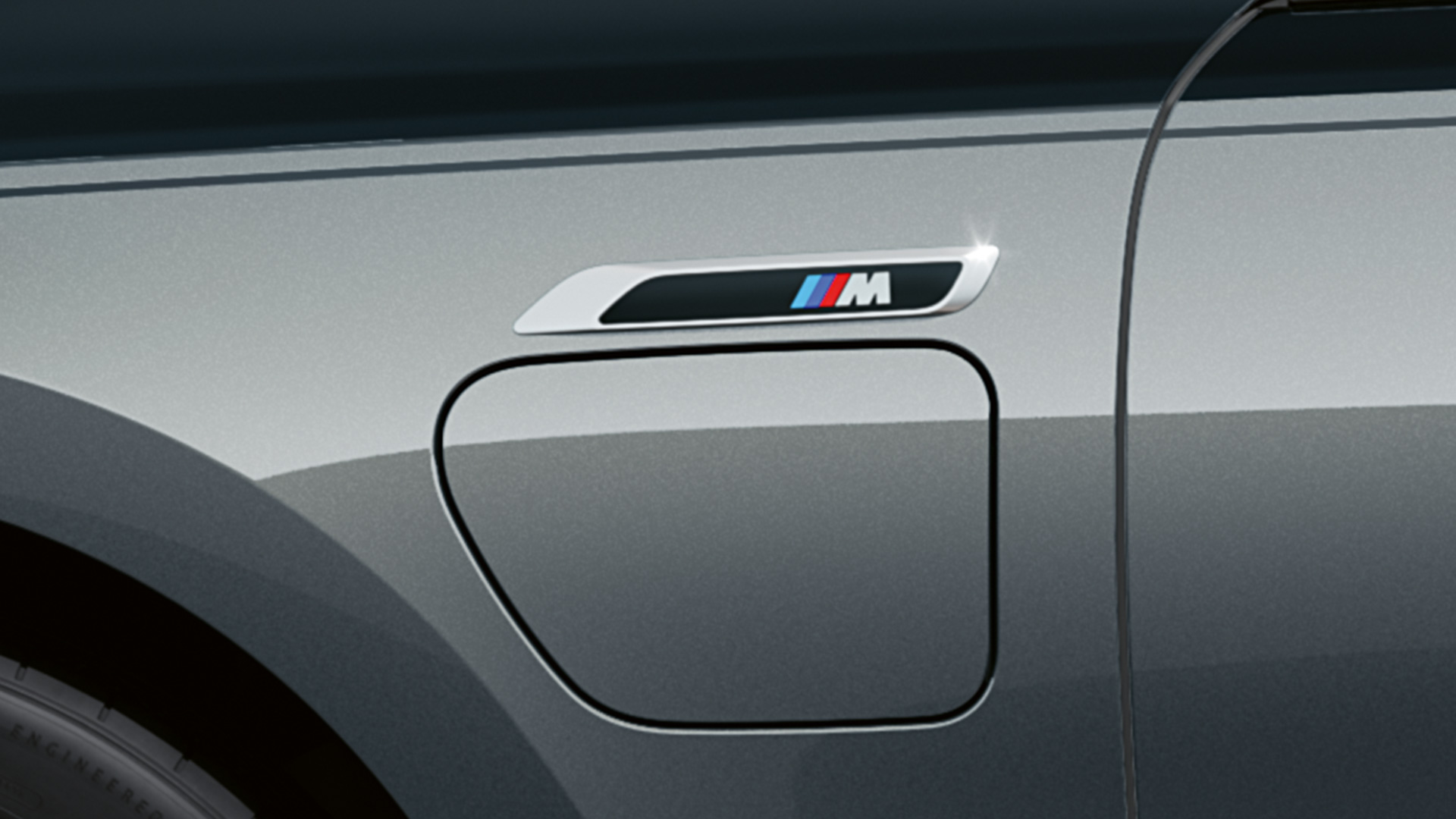 Логотип M BMW M760e xDrive G70 Логотип M на BMW M760e xDrive G70 с пакетом M Performance крупным планом, двухцветная окраска кузова BMW Individual (черный металлик Black Sapphire и серый металлик Dravite Grey), вид сбоку