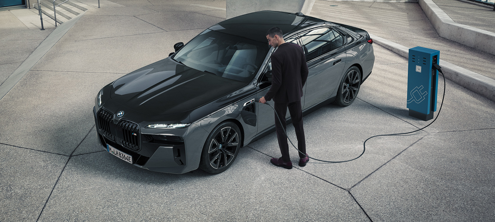 BMW M760e xDrive G70 с пакетом M Performance BMW M760e xDrive G70 с пакетом M Performance, двухцветная окраска кузова BMW Individual (черный металлик Black Sapphire и серый металлик Dravite Grey), вид спереди 3/4, мужчина держит зарядный кабель рядом с зарядной станцией