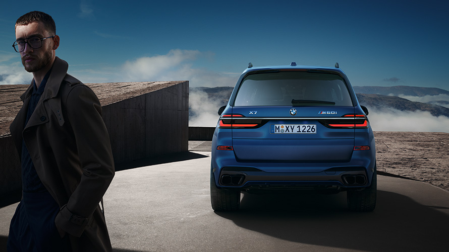 BMW X7 M60i xDrive G07 LCII шанақ бояуы қыраулы көк түс (Frozen Marina Bay Blue), тау пейзажы фонында модельмен түсірілген артынан көрінісі