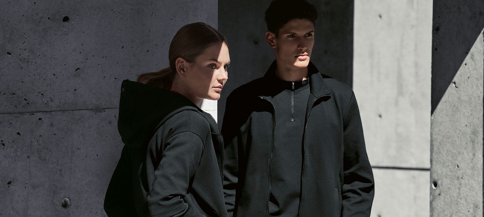 BMW M Sweat Jacket для женщин и BMW M Sweat Jacket для мужчин. Пара стоит в тени. Женщина представляет женскую куртку BMW M Sweat Jacket, а мужчина - мужскую куртку BMW M Sweat Jacket.