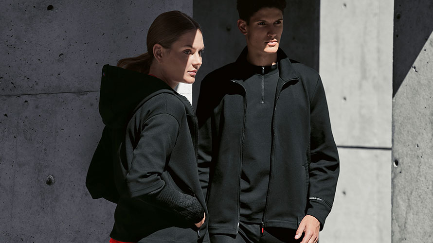 Пара стоит в тени. Женщина представляет женскую куртку BMW M Sweat Jacket, а мужчина - мужскую куртку BMW M Sweat Jacket.