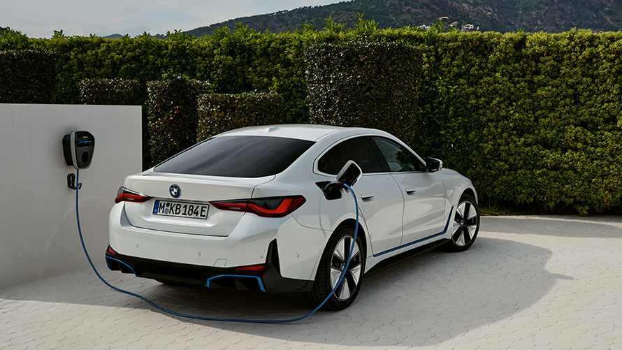 BMW i4 G26 BEV подключенный к BMW Wallbox Gen 3, вид сзади с поворотом в три четверти BMW i4 G26 BEV на фоне живой изгороди, подключенный к BMW Wallbox Gen 3, вид сзади с поворотом в три четверти